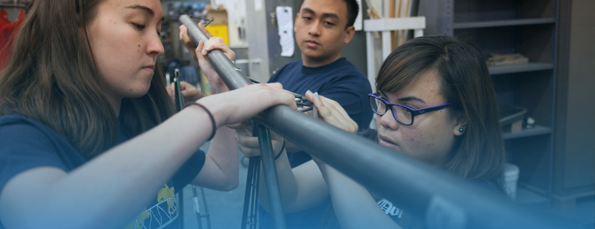 Three people working together on a long pipe in a cluttered lab, focused expressions