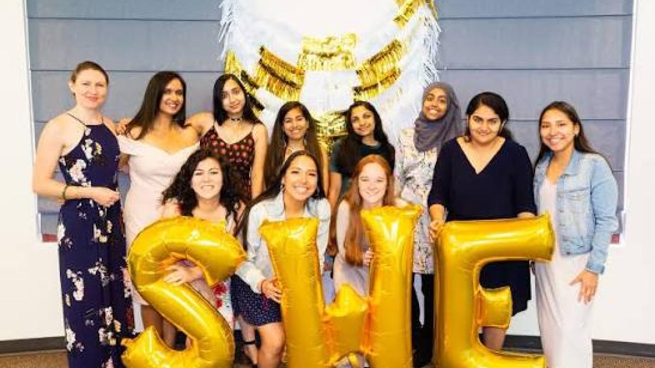 Eleven women smiling under white and gold balloon arch, holding gold SHE balloons