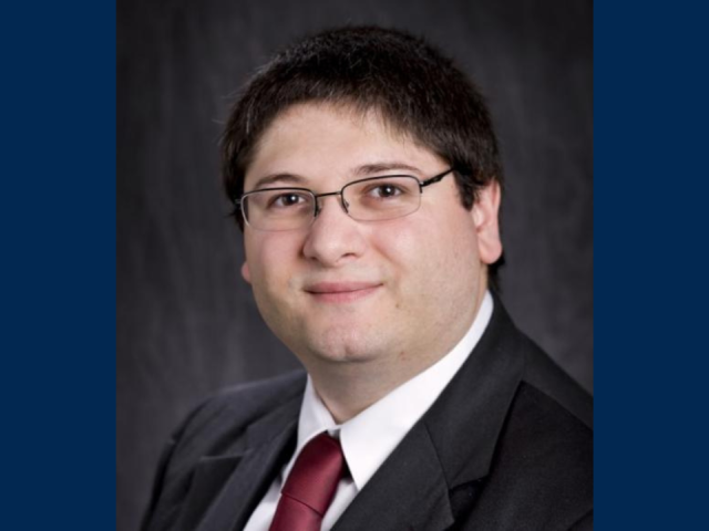 Studio headshot photo of a Barbato in suit and glasses against a dark backdrop