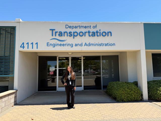 Person in dark suit standing outside Department of Transportation entrance, white facade, blue sign