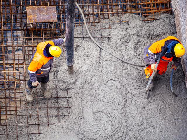 Two construction workers in yellow helmets pouring and leveling concrete over rebar, top-down.