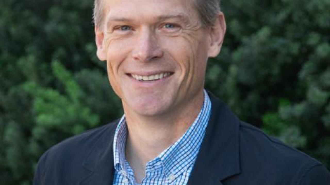 Photograph of professor Jason DeJong smiling and standing in front of some bushes