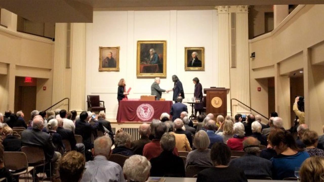 Image of a room filled with seated crowd watching a stage with two people on the left facing two people on the right. The center two people shaking hands behind a podium. Portraits of historic men hang on the wall behind them. 