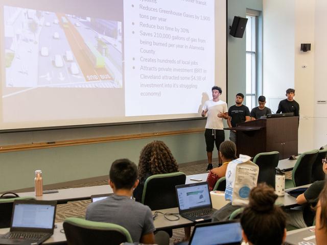 a group of male students standing behind a podium at the front of a classroom, presenting in front of their classmates. 