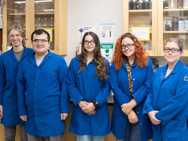 4 graduate students standing with hands crossed, all in blue lab coats, next to professor chris cappa. 