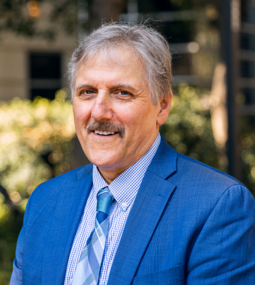 Photo of white man with gray hair and mustache outside wearing blue suit jacket, light blue shirt, and blue striped tie.