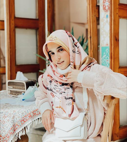 Hanieh sitting wearing a hijab at an indoor dining table, arm over chair.