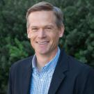 Photograph of professor Jason DeJong smiling and standing in front of some bushes